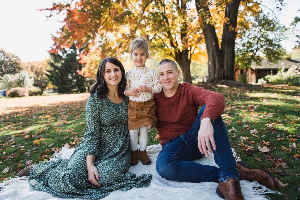 Family of three smiling, posing on a blanket in a park, autumn foliage.