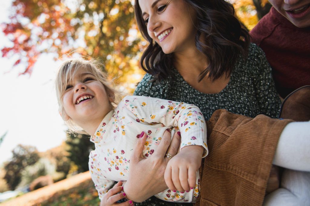 Smiling child being held, with a smiling adult, fall foliage in background.
