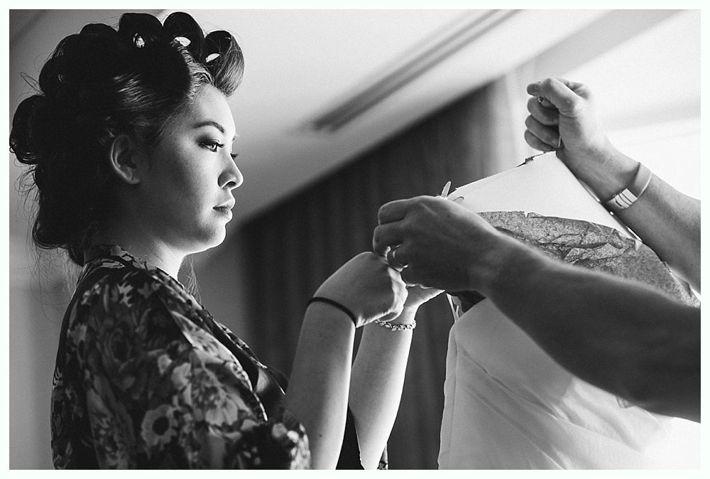 Woman with curlers gazes at dress being fastened; soft light in room.