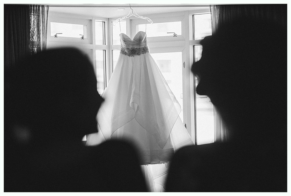 Wedding dress hanging in front of a window, silhouetted people looking at it.