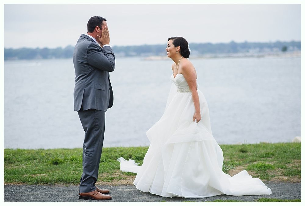Groom sees bride in her wedding dress, covering his face in surprise, waterfront setting.