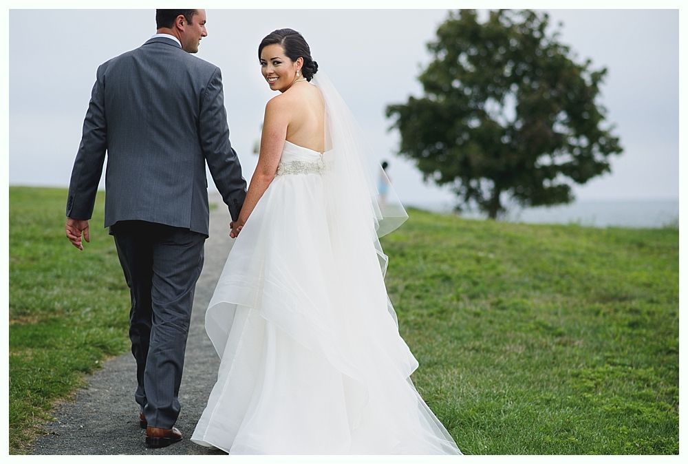Couple in wedding attire walk on a path, bride looks over shoulder, outdoor setting.