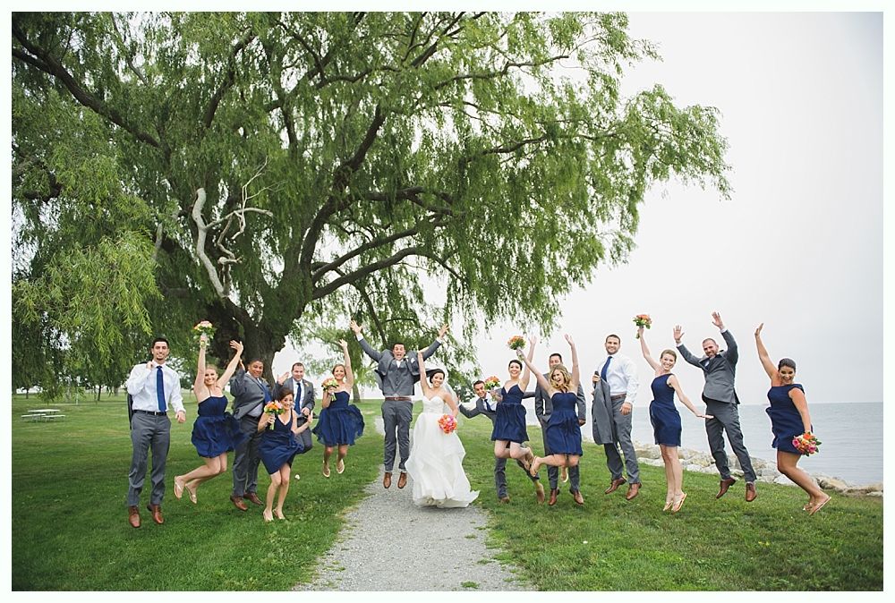 Wedding party jumping joyfully on a grassy path near a lake, under a large tree.