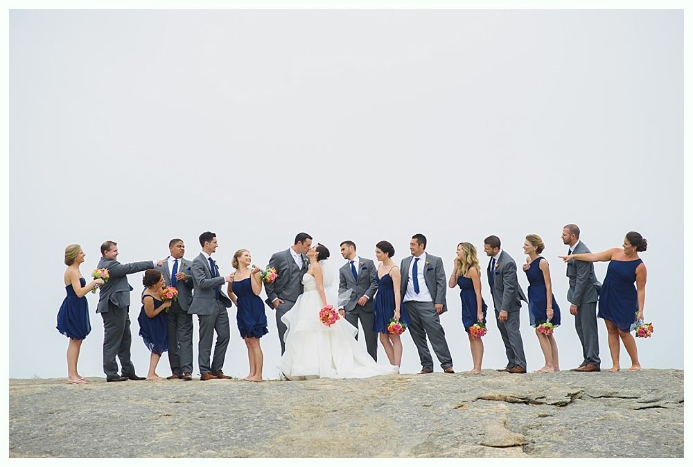 Wedding party on a rocky outcropping; bride and groom kiss with bridesmaids in blue dresses and groomsmen in gray suits.