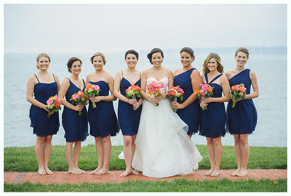Bridesmaids in navy dresses with the bride in white, holding bouquets, pose by the water.