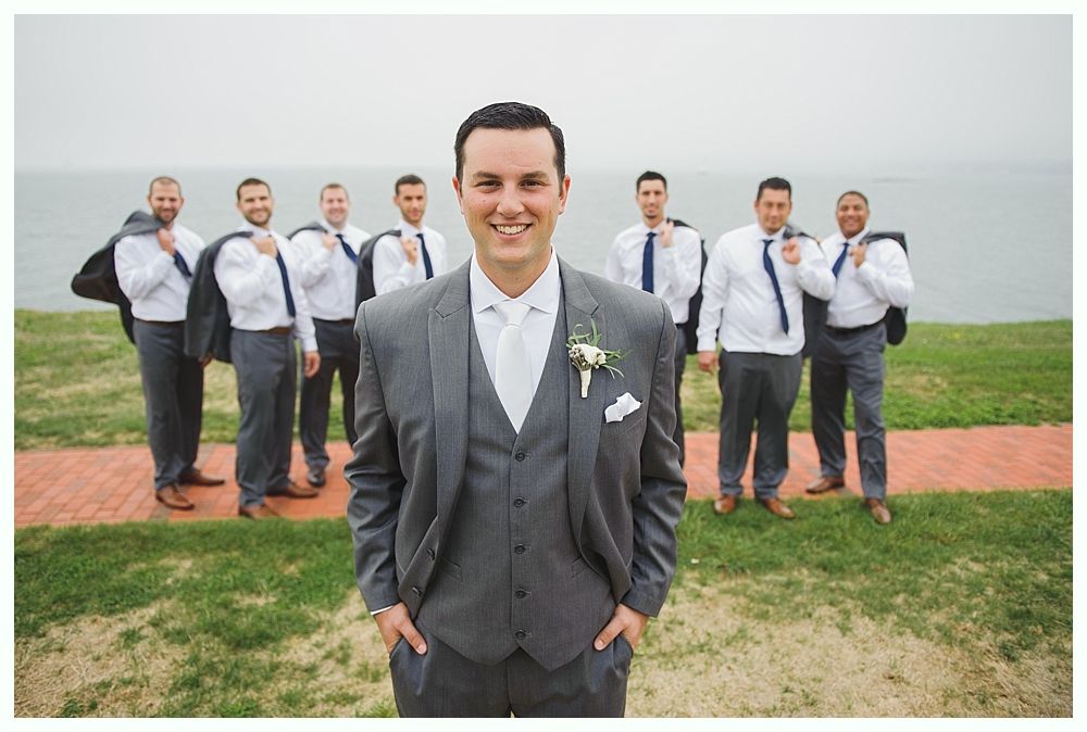 Groom in gray suit smiles, hands in pockets; groomsmen in white shirts, ties, jackets over shoulders, standing behind him near water.