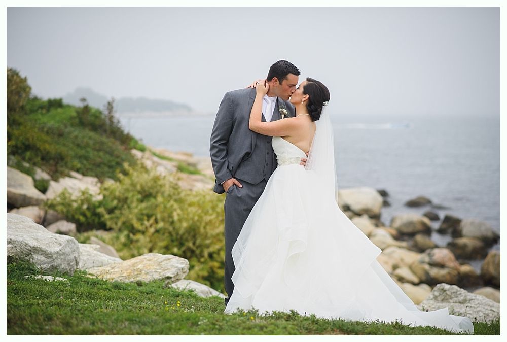 Bride and groom kissing outdoors. The bride wears a white strapless gown and veil, while the groom wears a gray suit. They stand by the ocean.