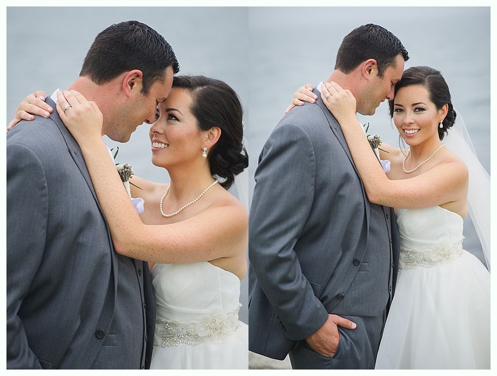 Wedding couple in embrace, gray suit, white dress, ocean backdrop, smiles.