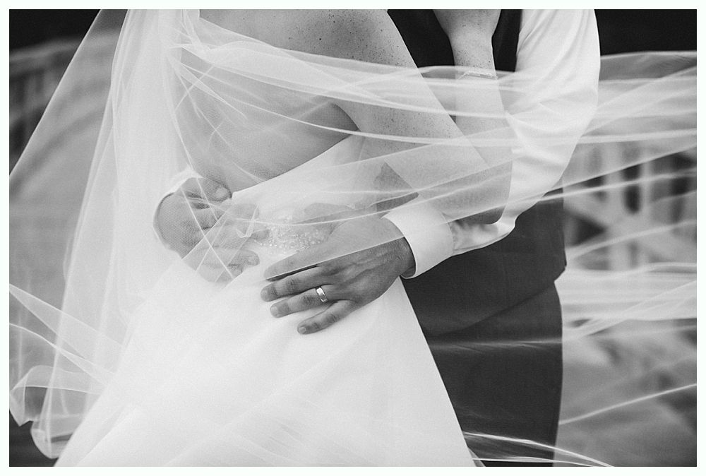 Bride and groom embrace, veil swirling around them, rings visible. Black and white.