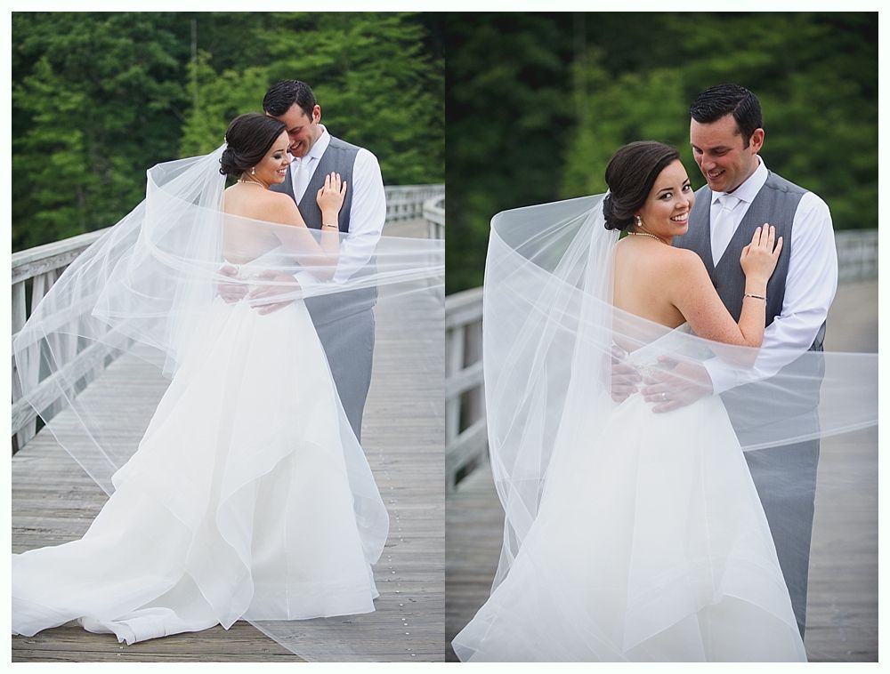 Bride and groom embracing on wooden bridge. Bride wears white gown and veil, groom in gray vest and shirt.