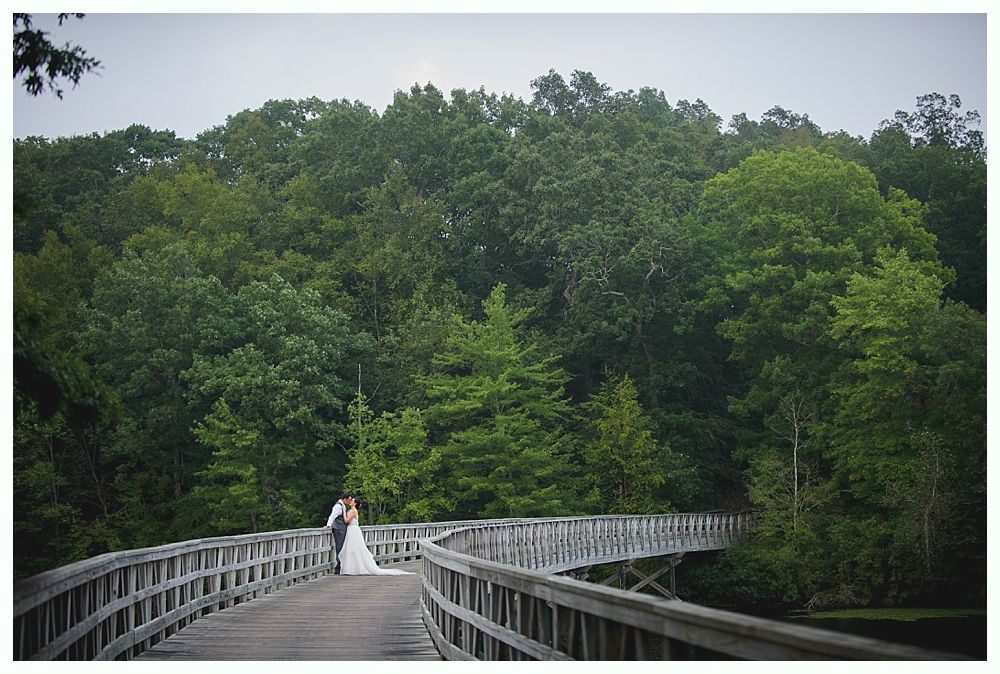 Couple on a wooden bridge, embracing in front of a lush green forest on a cloudy day.