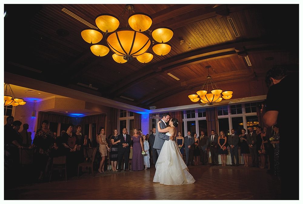 Couple dances at a wedding reception, lit by chandeliers. Guests watch on a wooden dance floor.
