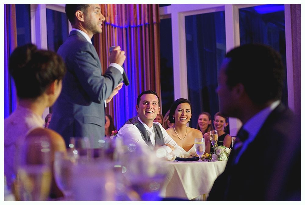 Man giving speech at a wedding, couple smiling, other guests watching, elegant setting.