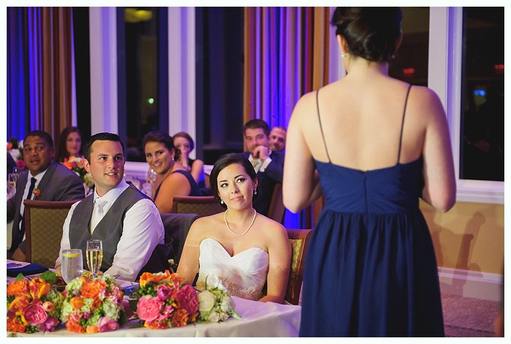 Wedding reception: bridesmaid giving a speech, bride and groom seated, guests listening.