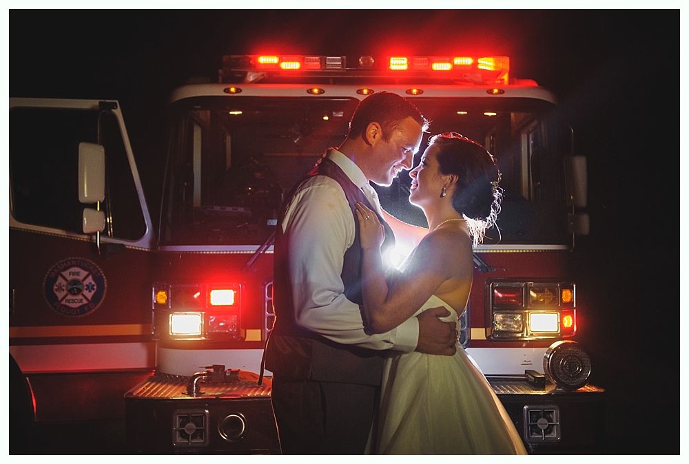 Couple in wedding attire embrace in front of a fire truck, lit by flashing lights.