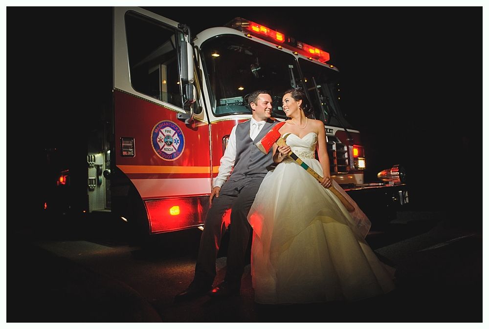 Couple in wedding attire pose by a fire truck at night; emergency lights are on.