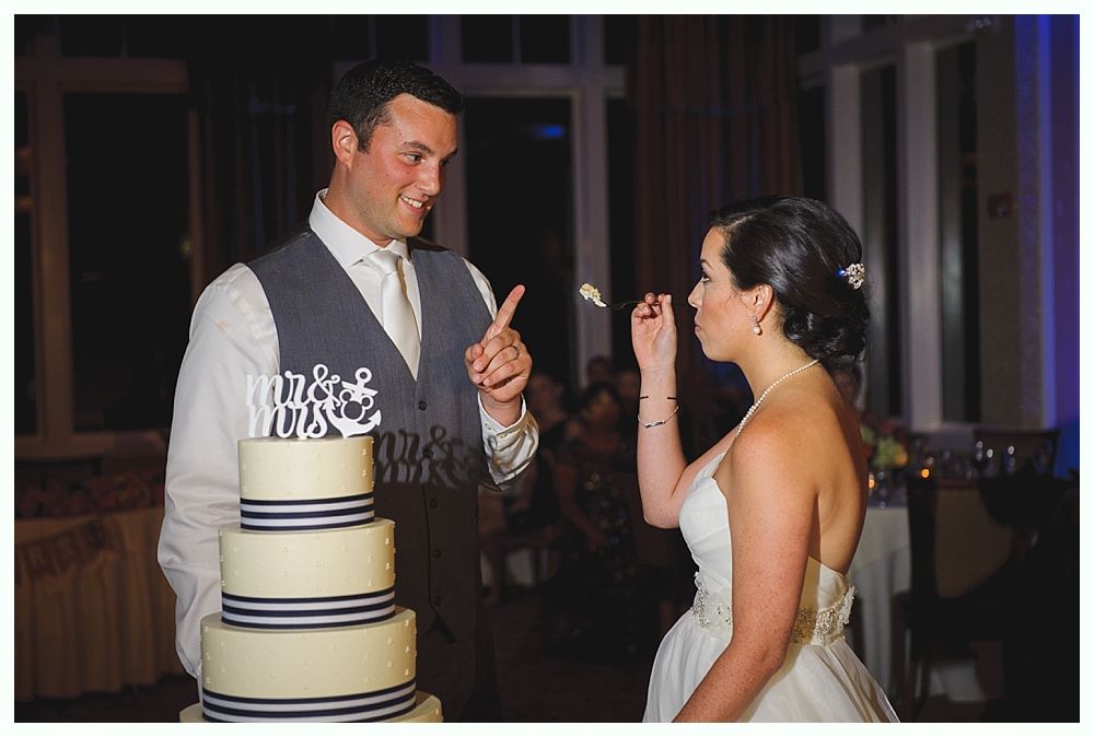 Couple at wedding cake, man gesturing as woman takes bite. Cake has navy stripes, 