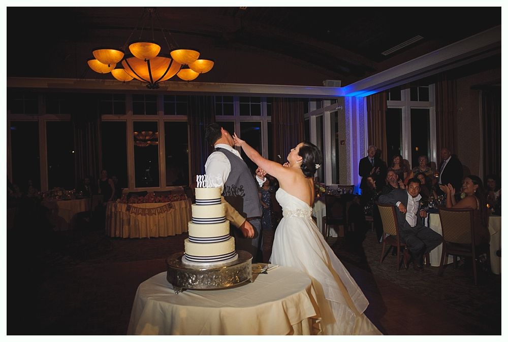 Bride feeds groom wedding cake, they are in a dimly lit reception hall. Cake has multiple tiers, guests watching.