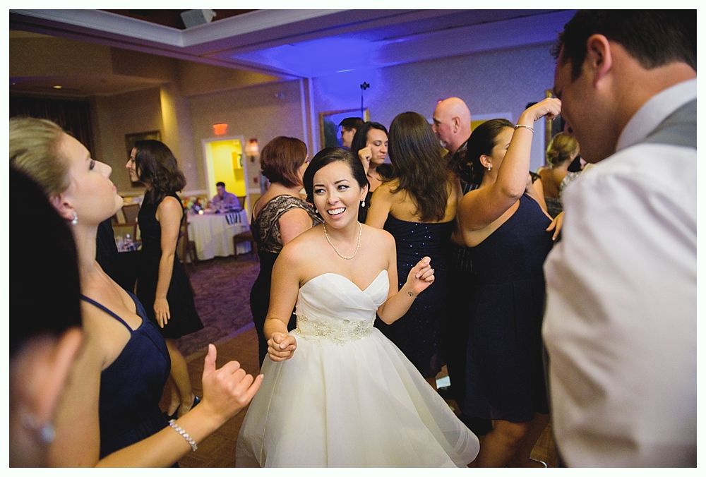Bride dancing joyfully at a wedding reception; surrounded by guests. Ballroom lit with blue and gold hues.