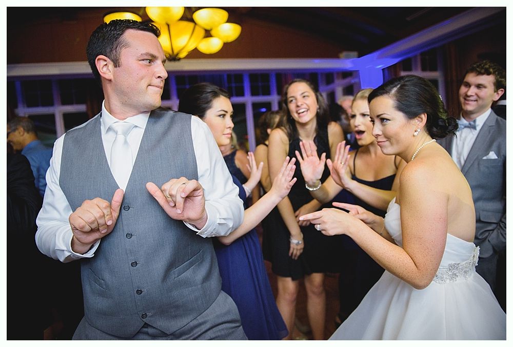 People dancing at a wedding reception. The groom dances enthusiastically; the bride and other guests watch, laughing.