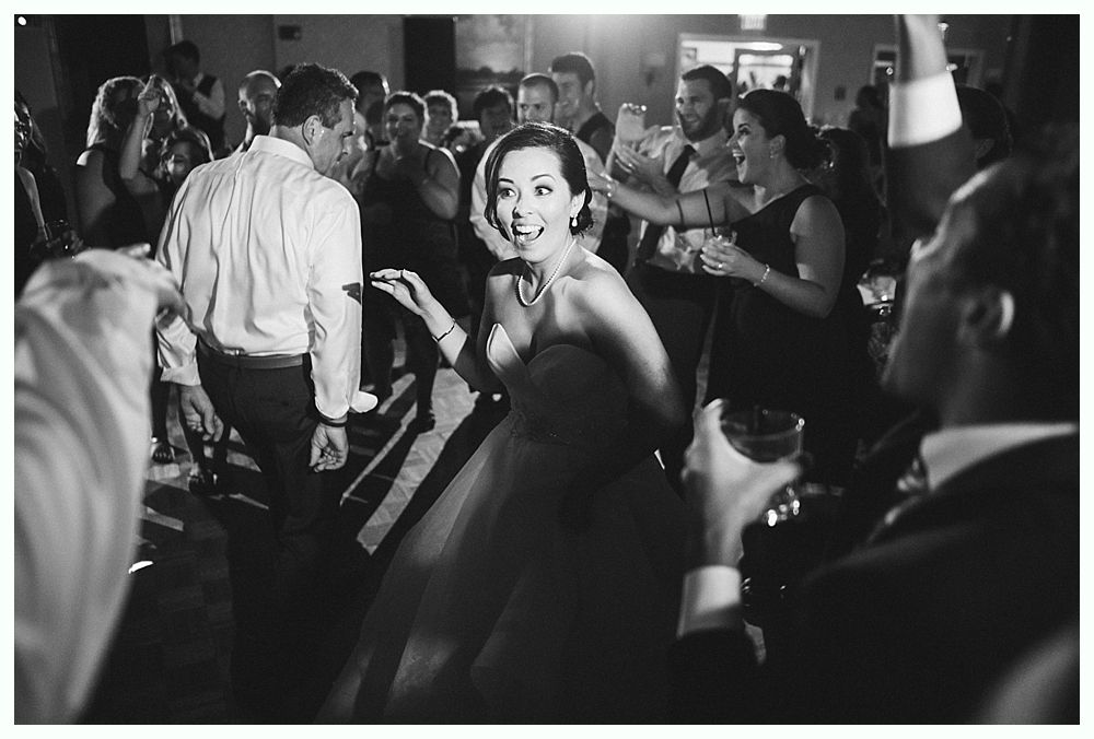 Woman in a strapless gown dances, smiling, surrounded by people at a dimly lit event.