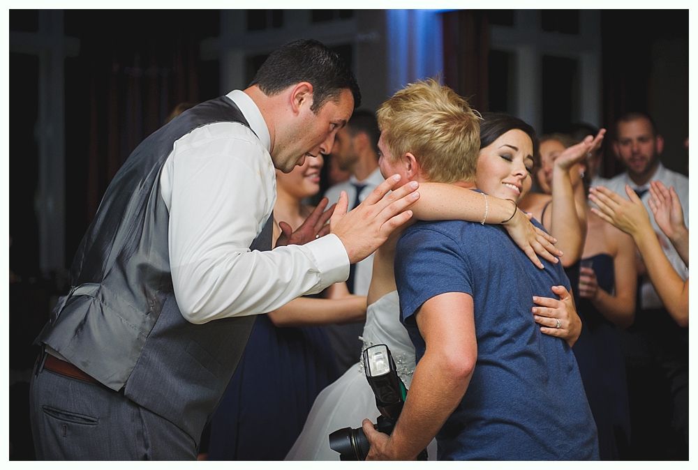 Wedding celebration with bride embraced by two people, one holding a camera, people cheering in the background.