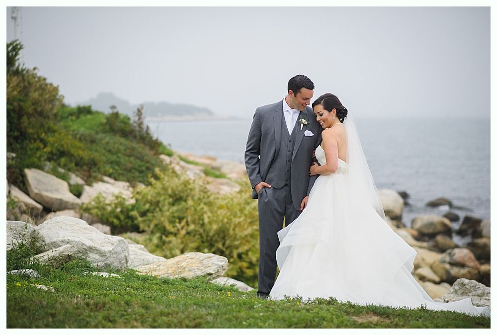 Bride and groom pose on grassy shore by ocean on a cloudy day. The groom wears a grey suit, and the bride has on a white dress.