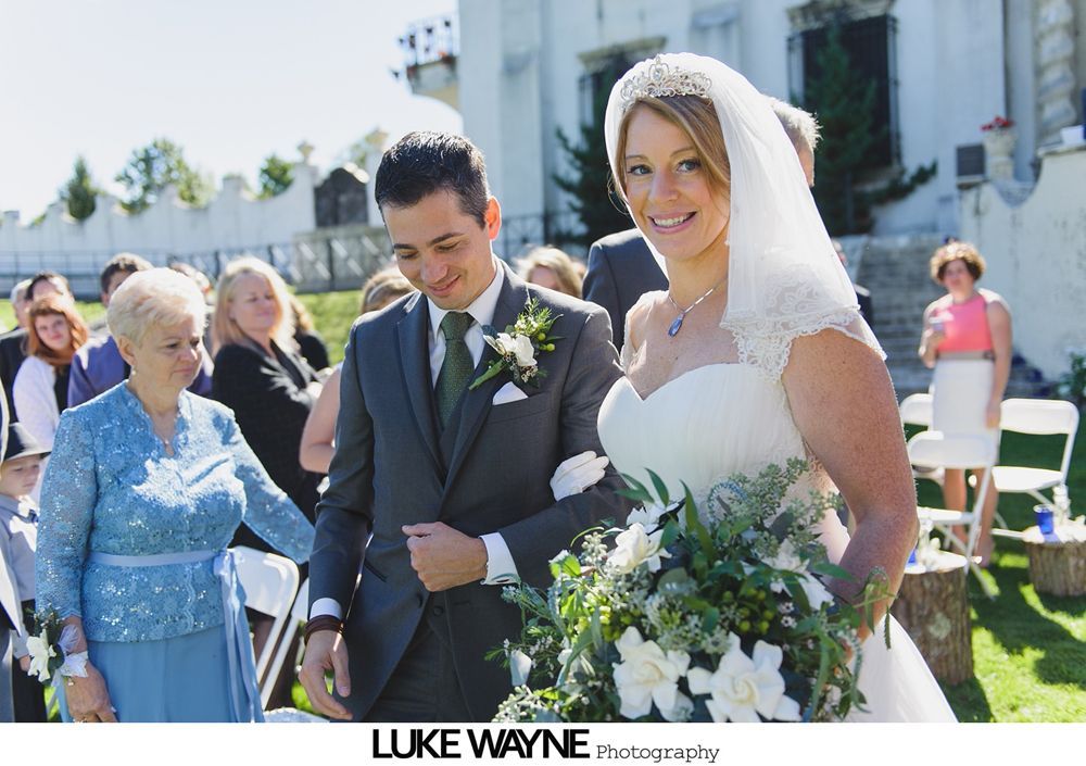 Bride and groom embrace, autumn leaves in background. Groom kisses bride's cheek. Sunlight creates yellow glow.