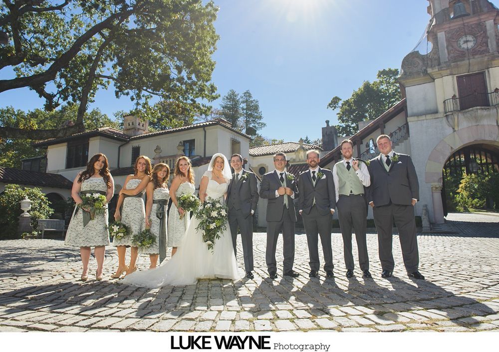 Wedding ceremony with couple, wedding party, and guests outdoors on a grassy lawn with trees and blue sky.