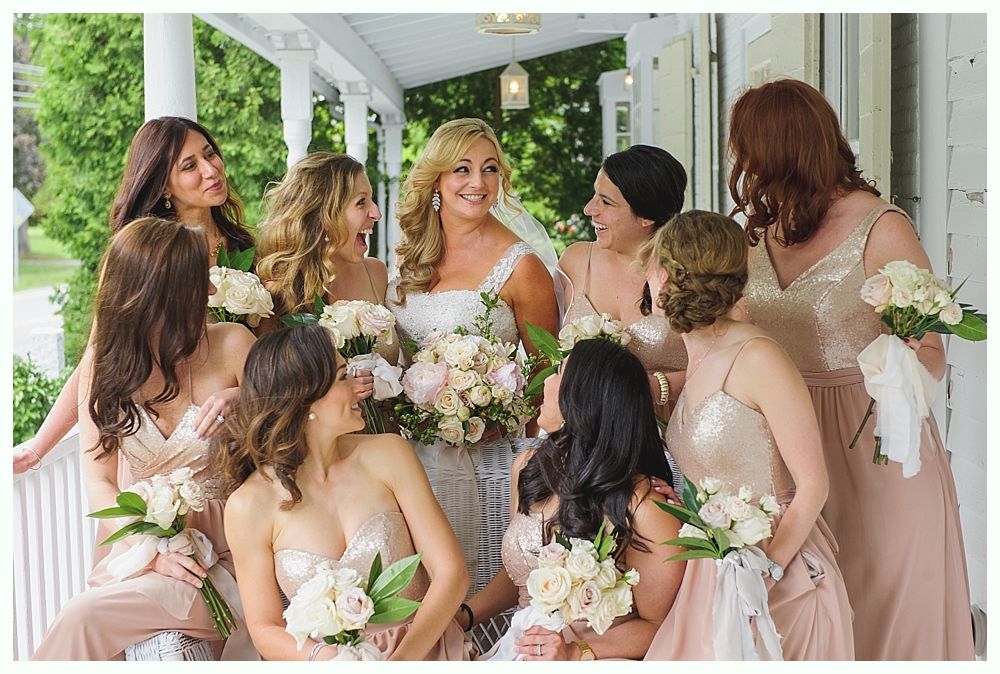 Bride surrounded by bridesmaids holding bouquets, all in neutral dresses, on a porch.