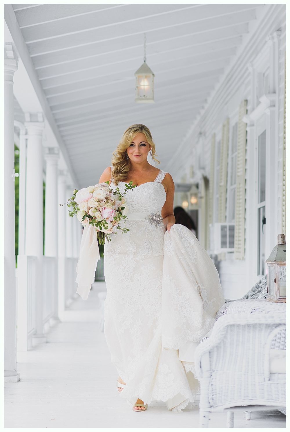 Bride in white lace wedding dress walks on a white porch, holding bouquet.