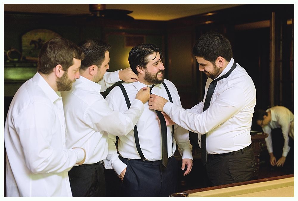 Four men in white shirts and black ties helping each other adjust ties and suspenders in a dark room.