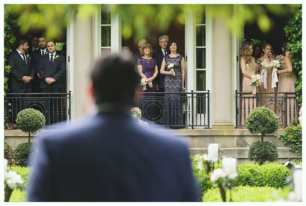 Groom looking at family on balcony. Ceremony setting with floral decor and greenery. Formal attire, expectant expressions.