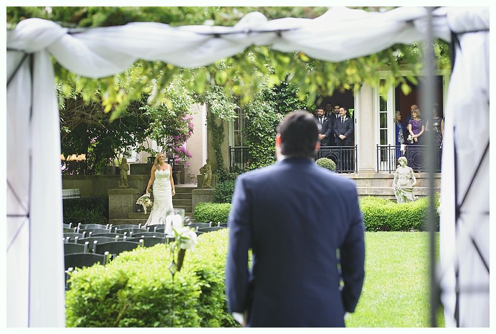 Groom awaits bride at outdoor wedding ceremony; white archway frames the view.