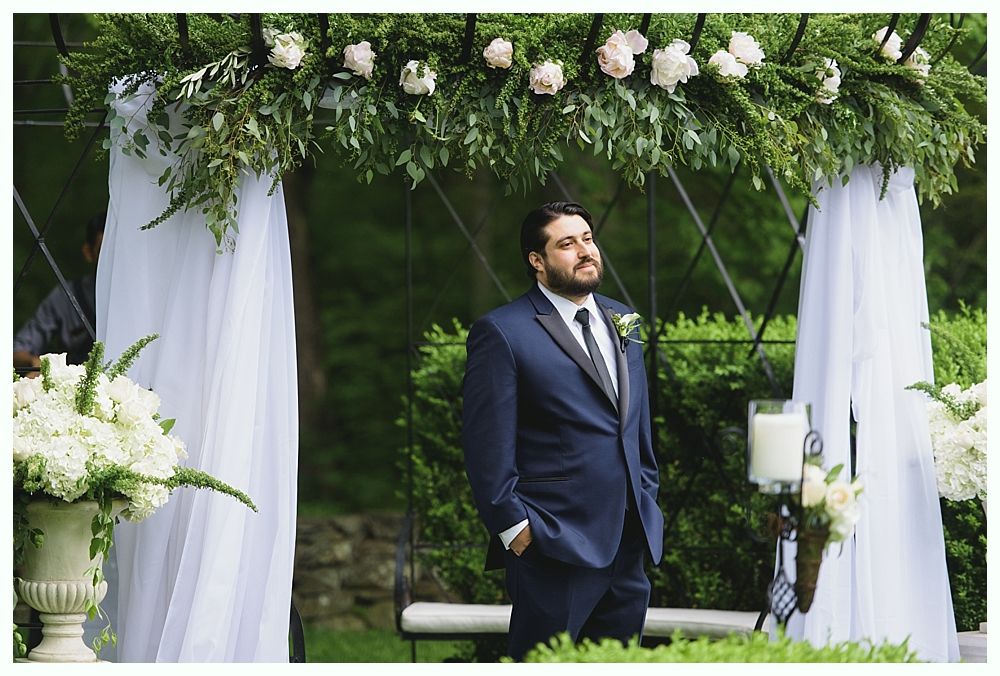 Man in a blue suit stands under a floral archway, awaiting a wedding ceremony.