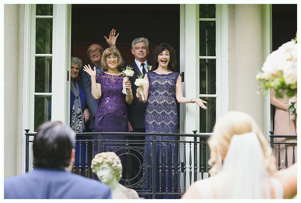 Family waves from a balcony to a bride holding flowers; building exterior, sunny day.