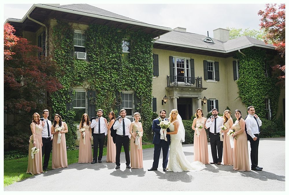 Wedding party posing in front of a large house covered in ivy. Bride and groom centered. Peach bridesmaid dresses.