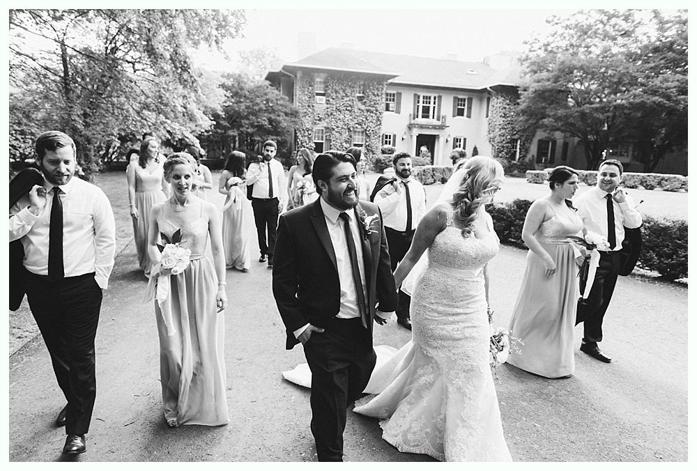 Wedding party walking on a path toward a large house, bride in a gown, groom in a suit, others in formal wear.