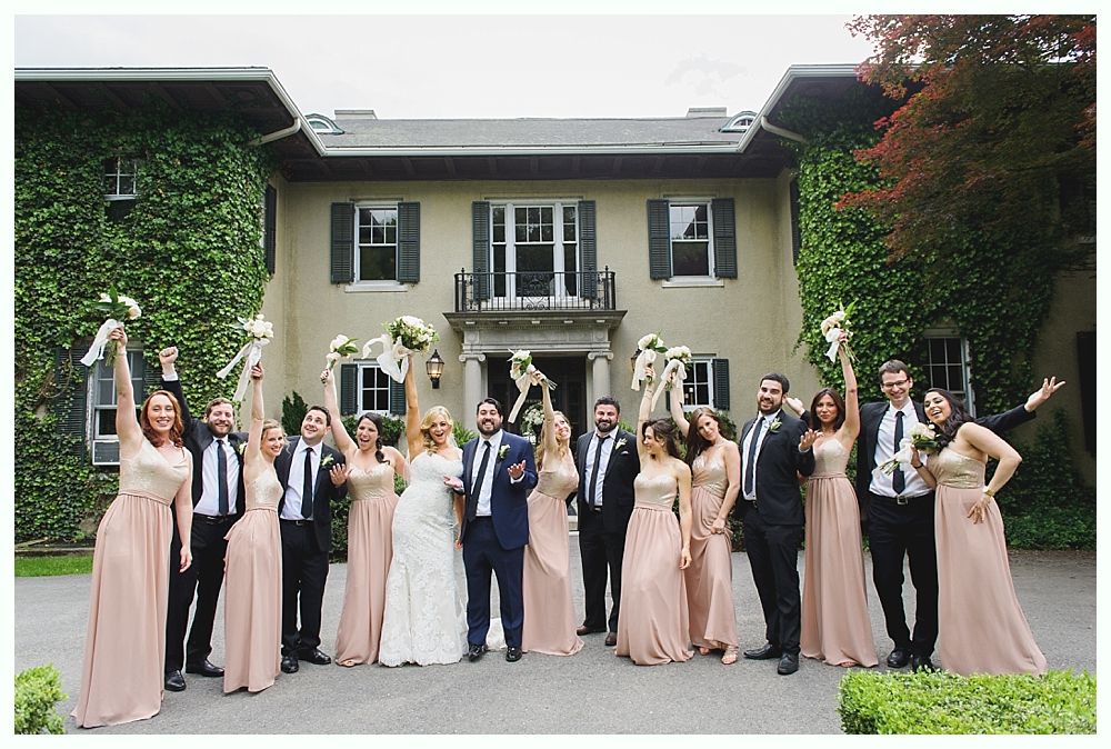 Wedding party in front of a large house, raising bouquets. Bride and groom centered; bridesmaids in peach, groomsmen in black.