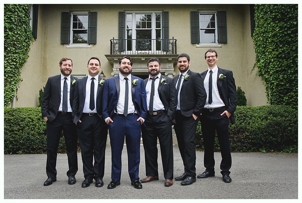 Six men in suits pose outside a building. One in blue suit stands in the center.