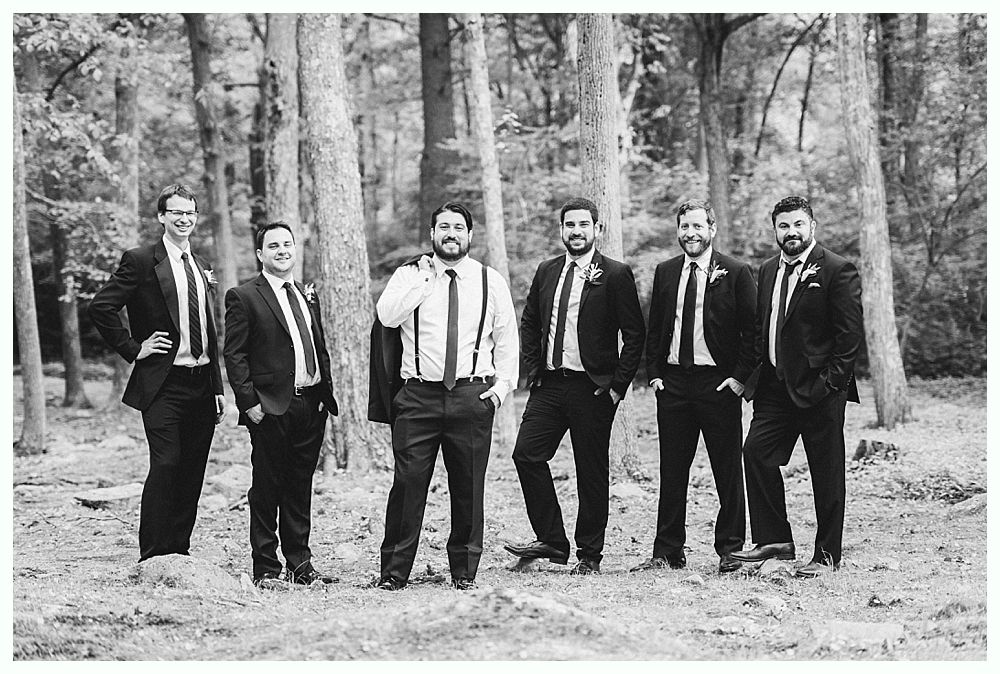 Men in suits pose outdoors among trees. One holds his jacket.