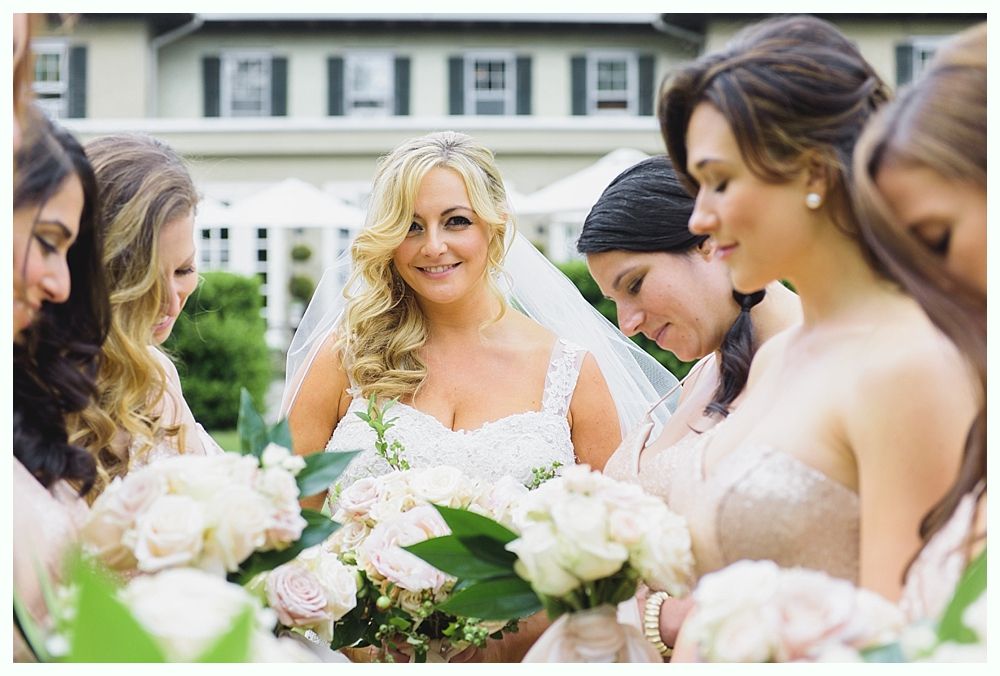 Bride in white dress smiles surrounded by bridesmaids in pink dresses, holding bouquets outdoors.