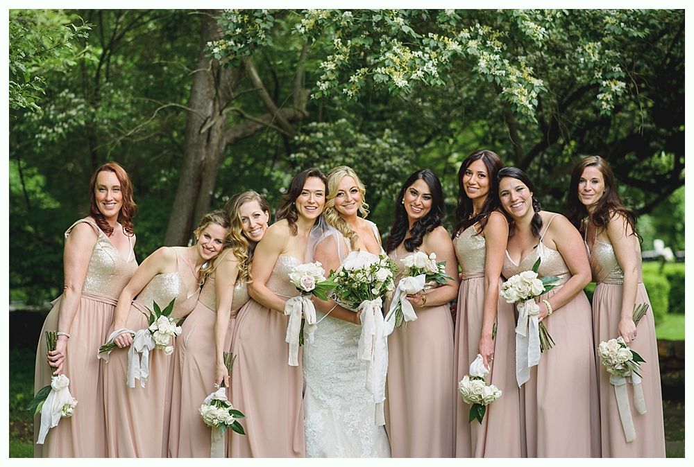 Bridesmaids in blush dresses pose with bride in front of greenery.