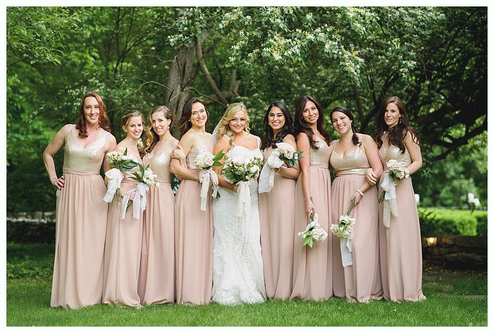 Bridesmaids in blush dresses pose with bride in white dress, holding bouquets. Outdoors, green foliage.