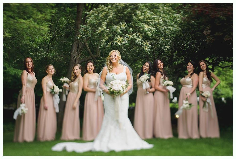 Bride and bridesmaids in blush gowns with bouquets pose on a lawn. Lush green foliage forms a backdrop.