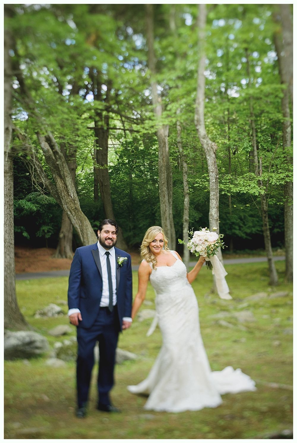 A smiling bride, in a white dress, playfully holds a bouquet while the groom stands beside her in a forest.