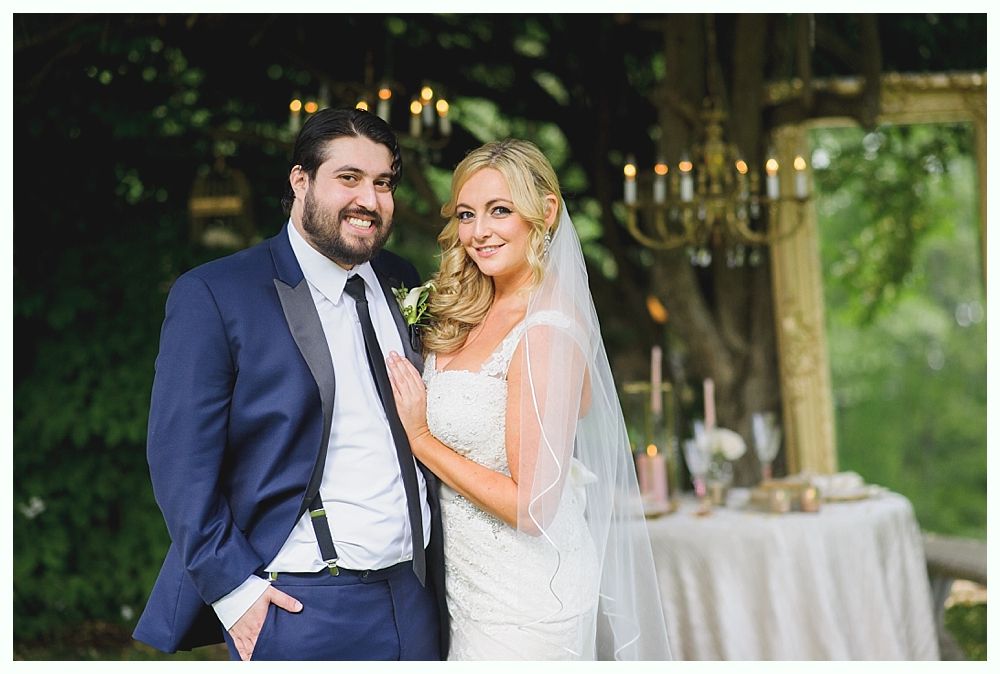 Bride and groom smiling at camera outdoors; man in blue suit, woman in white dress.