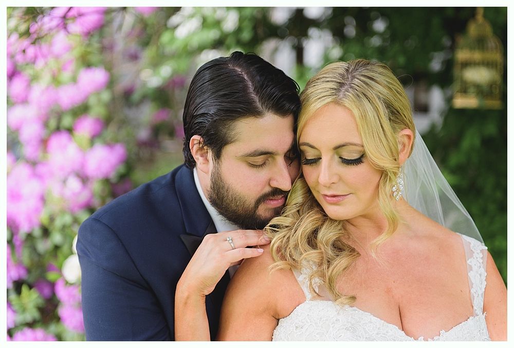 Bride and groom embrace outdoors; she in white dress and veil, he in blue suit, pink flowers in background.
