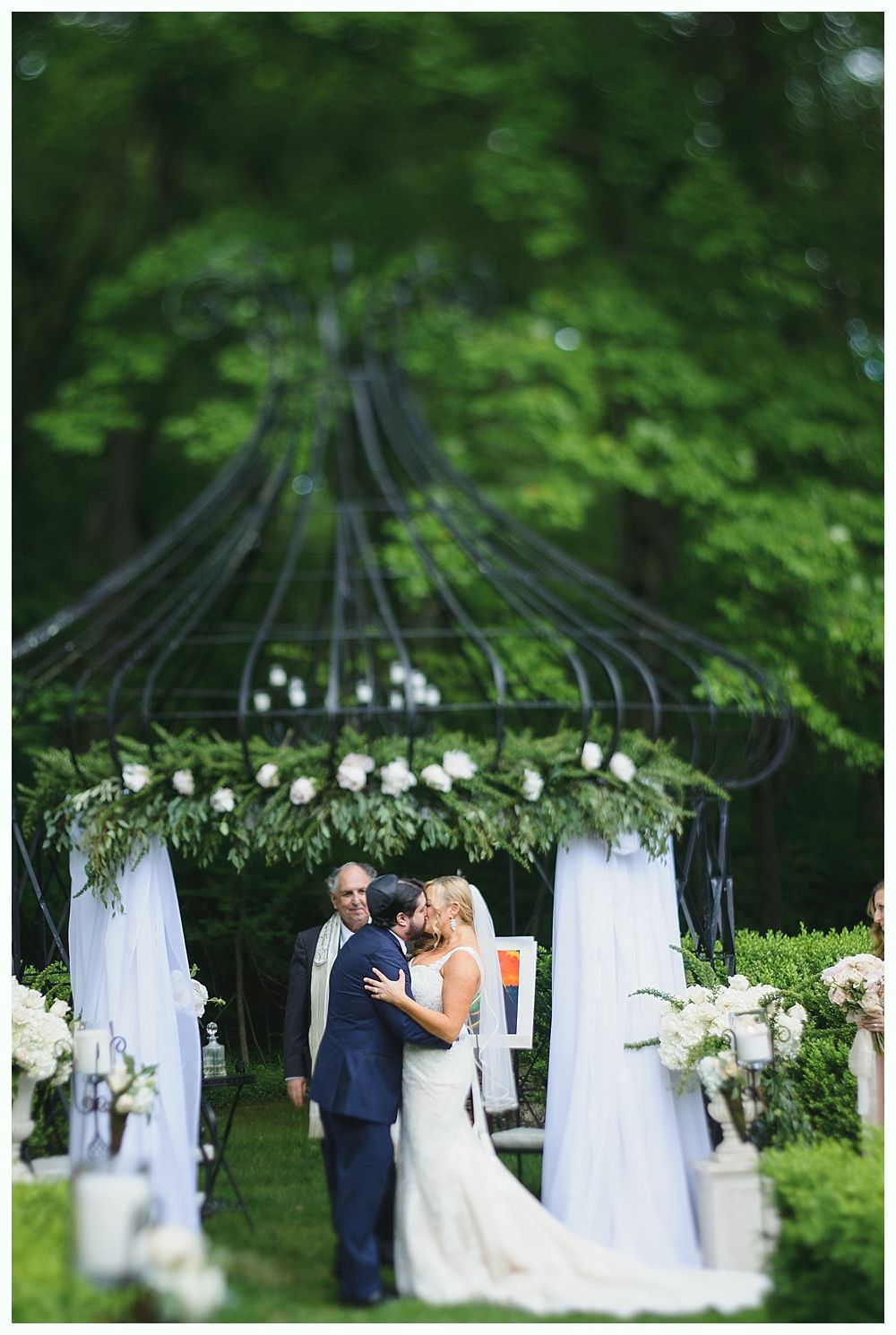 Bride and groom embrace under a gazebo during wedding ceremony in a garden setting.