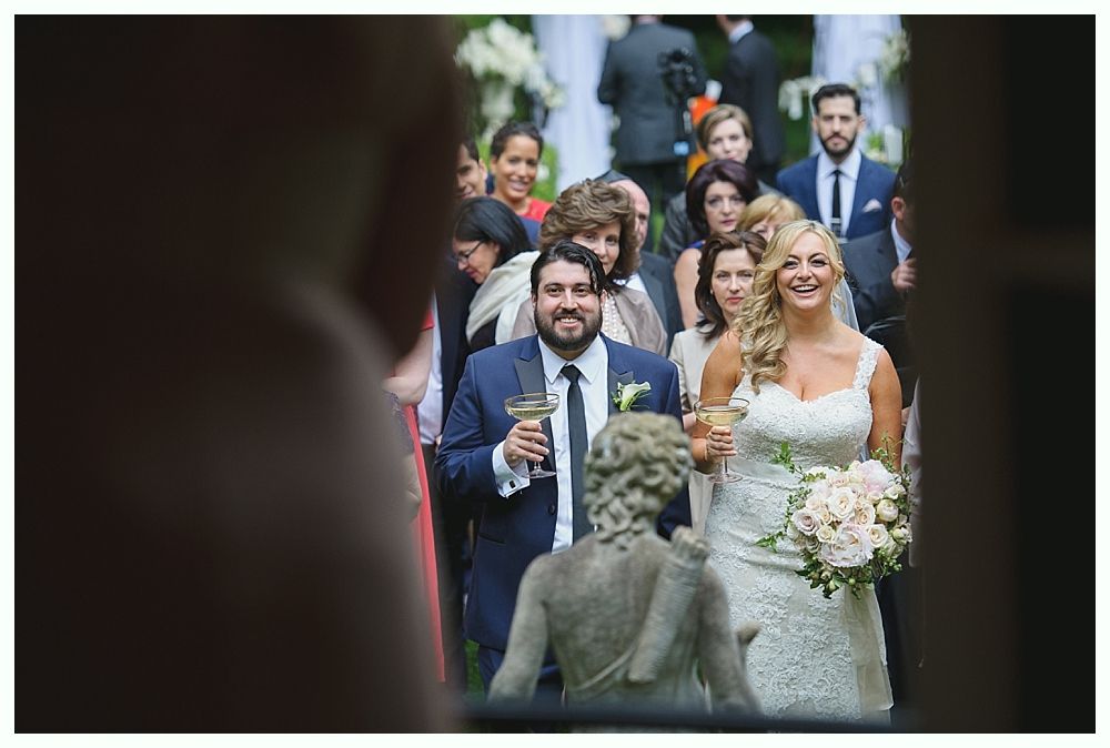 Newlyweds and wedding party walking, holding drinks, smiling. Outdoor, sunny setting.